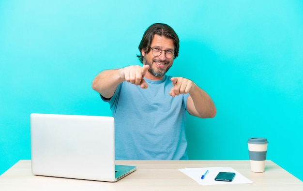 Senior dutch man in a table with a laptop isolated on blue background pointing to the front and smiling