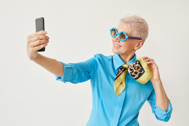 흰색 배경에 대해 그녀의 휴대 전화에 카메라에 포즈 선글라스 수석 우아한 여자
