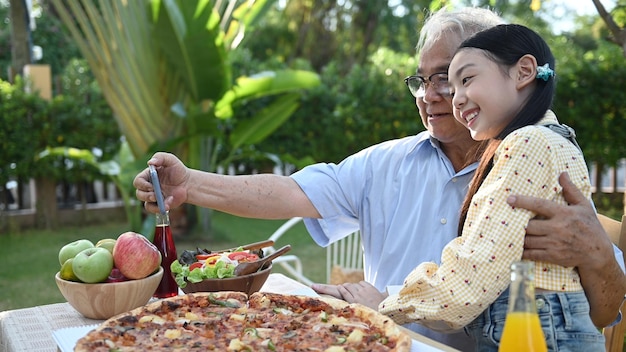 Senior en kleindochter met pizza in de tuin thuis. Pensioenleeftijd levensstijl met familie op zomervakantie.