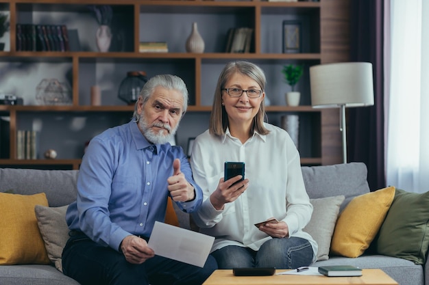 Senior familiepaar grijsharige oudere betaaldiensten voeren gegevensdocumenten in met behulp van applicatie mobiele smartphone creditcard Gepensioneerden ouderen thuis nutsrekeningen cheques gebruiken computer slimme telefoon