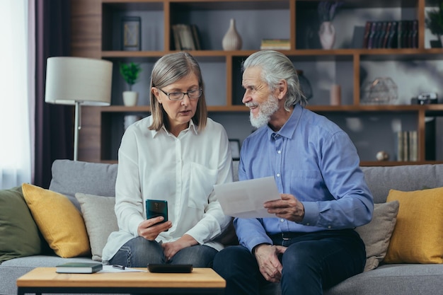 Senior family couple gray haired old man and woman pay for\
services enter data documents using application mobile smartphone\
retirees at home utility bills checks use computer smart phone