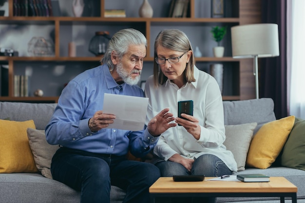 Senior family couple gray haired old man and woman pay for\
services enter data documents using application mobile smartphone\
retirees at home utility bills checks use computer smart phone