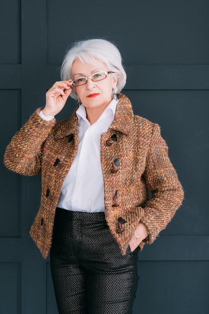シニアファッションスタイリストの肖像画。トレンディなワードローブのパーソナルコンサルタント。眼鏡をかけて手でポーズをとる自信のある老婦人。