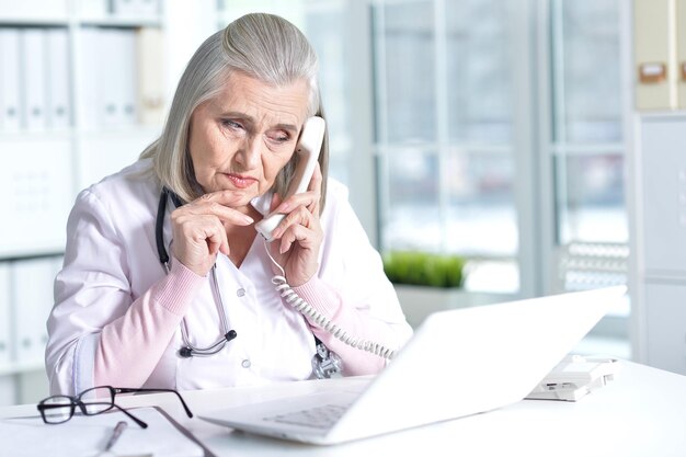 Senior female doctor using laptop and talking on the phone