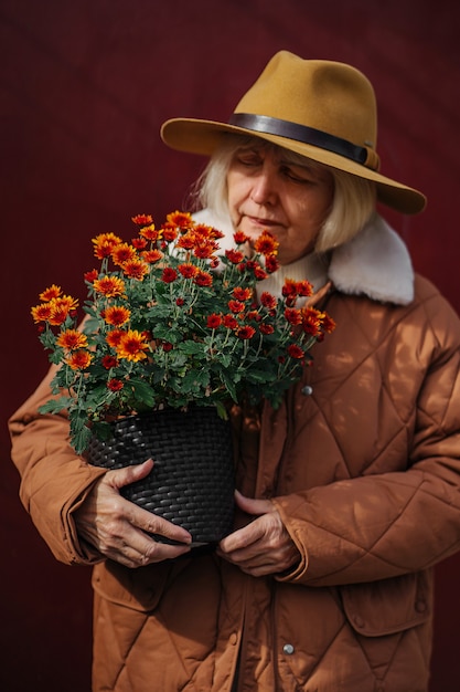 ワインの背景に対して鉢植えの花の束を検査するアウターウェアのシニア女性庭師。