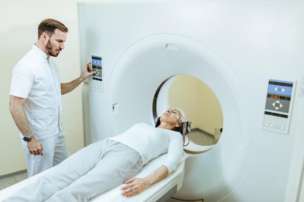 Пожилая пациентка проходит компьютерную томографию, пока радиолог наблюдает за процедурой