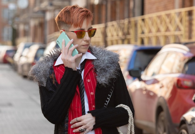 빨간색 슈트와 선글라스를 입은 고령 여성이 거리에서 스마트폰으로 대화하는 동안 미소 짓고 있습니다.