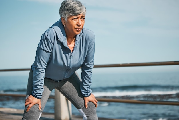 Senior fitness en vermoeide vrouw op het strand tijdens pauze van trainingstraining of ochtend cardio-run in de natuur Sportvermoeidheid en oudere vrouwelijke hardloper stoppen om te ademen tijdens oceaantraining of prestatie