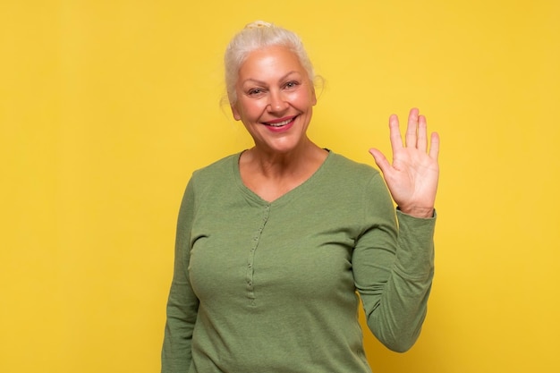 Senior friendly looking woman smiling happily saying Hello Hi or Bye waving hand at camera