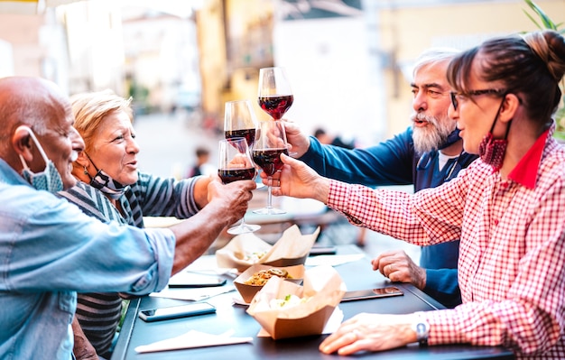 Senior friends toasting red wine at winery bar dehor with open face mask