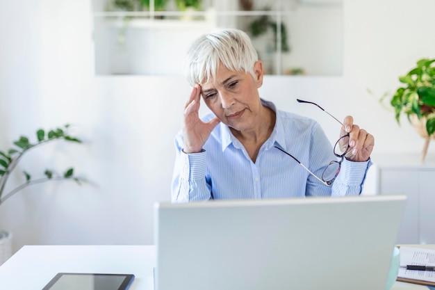 Senior gefrustreerde vrouw die vanuit thuiskantoor voor laptop werkt, hoofdpijn heeft. Volwassen vrouw met een pijnlijke gezichtsuitdrukking