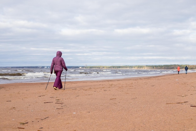 Senior gepensioneerde vrouw wandelen aan de kust op het zand Actieve fitness en gezonde levensstijl Vrouwelijke training op het strand Nordic walking met stokken Herfstseizoen aan de kust Sportief paar