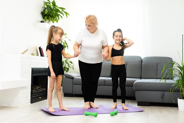 senior grandmother workout yoga withgranddaughters preschooler. They are sitting on mat at cozy home interior. Sport, parenthood and people concept.