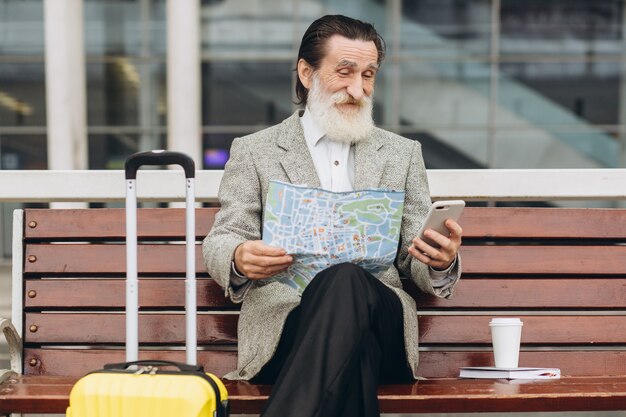 Senior uomo con la barba grigia si siede su una panchina con una valigia, guarda la mappa della città e il telefono all'edificio dell'aeroporto