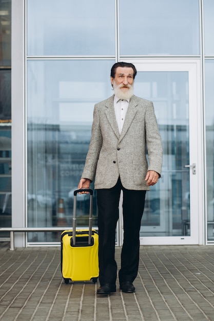 Senior gray-bearded man with carrying suitcase and city map is walking along the airport building. He is looking aside pensively. Copy space on right side