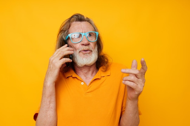 Senior greyhaired man wearing blue glasses yellow shirt talking on the phone isolated background