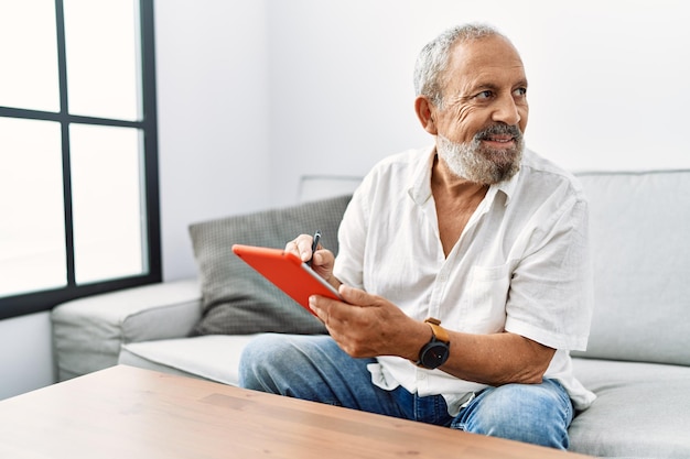 Senior grijsharige man glimlachend zelfverzekerd met behulp van touchpad thuis