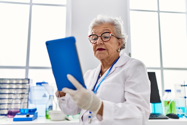 Senior grijsharige vrouw in wetenschapperuniform met touchpad in laboratorium