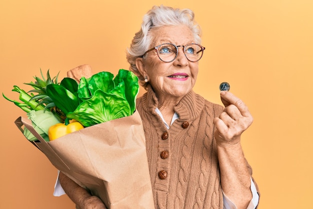 Senior grijsharige vrouw met boodschappen en 1 euromunt glimlachend opzij kijkend en denkend weg starend.