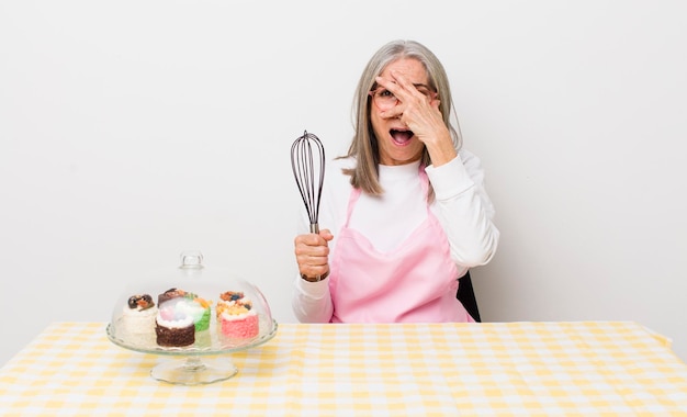Senior grijze haarvrouw die geschokt, bang of doodsbang het gezicht bedekt met handgemaakt gebakconcept