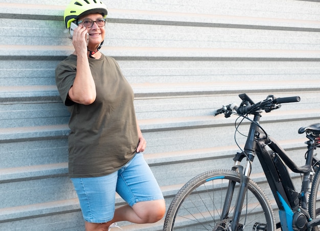 Senior grootmoeder met mobiele telefoon leunend tegen een metalen muur. Gezonde levensstijl met de elektrische fiets. Gele helm om te beschermen.