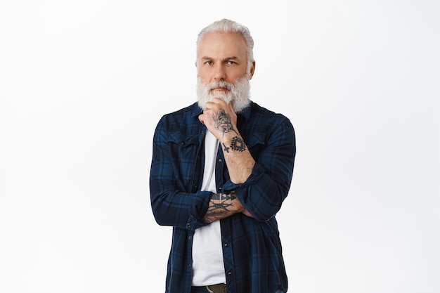 Senior guy with tattoos pondering decision, making choice, standing over white wall