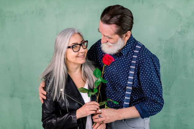 Senior handsome bearded man offering a rose to his partner, charming gray haired lady, standing together in front of green wall