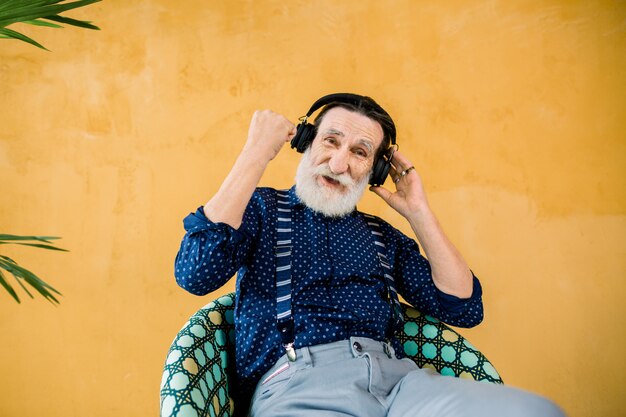 Uomo barbuto bello senior che indossa i vestiti alla moda che si siedono sulla sedia vicino alla parete gialla con la palma