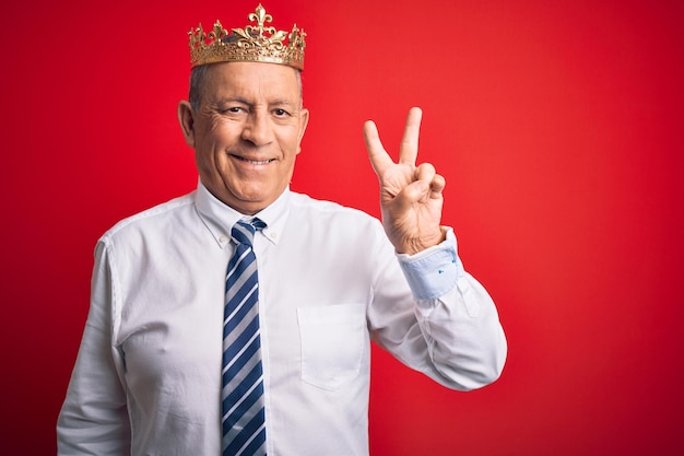 Старший красивый бизнесмен в королевской короне, стоящий на изолированном красном фоне, улыбающийся со счастливым лицом, подмигивающий в камеру, делая знак победы номер два