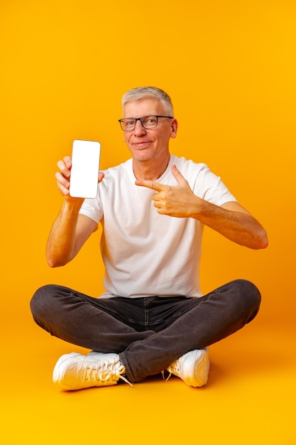 Senior handsome smiling man holding smartphone showing screen over yellow background