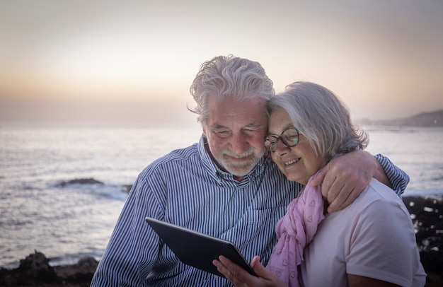 ビーチで夕暮れ時に座っている同じタブレットを見て楽しんでいるシニア幸せなカップル。テクノロジーを楽しんでいる退職者、海の地平線