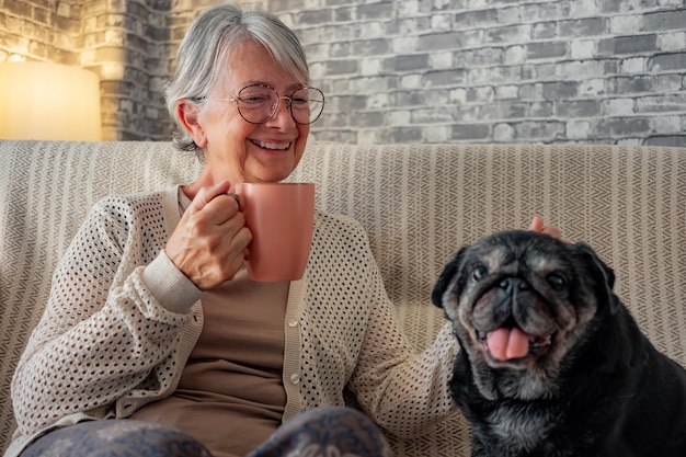 Senior happy woman sitting on sofa with her best friend an old black pug dog Elderly lady relax with a coffee cup in good company