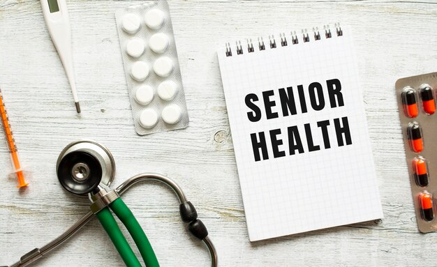 SENIOR HEALTH is written in a notebook on a white table next to pills and a stethoscope.