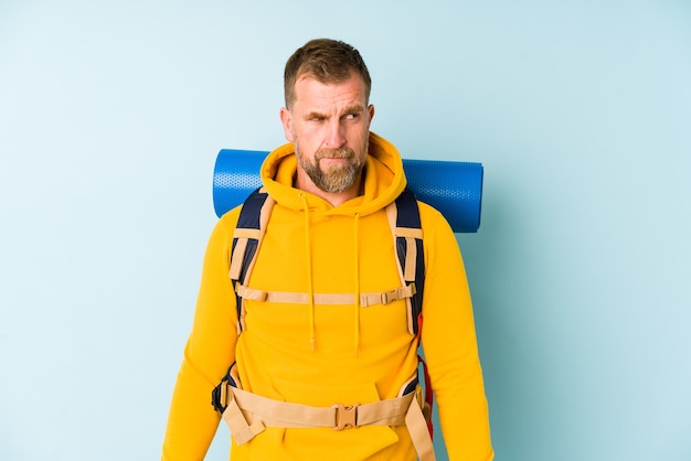 Senior hiker man isolated on blue wall confused, feels doubtful and unsure.