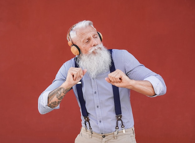 Senior hipster man danst buiten terwijl hij naar muziek luistert met een draadloze koptelefoon met rode achtergrond