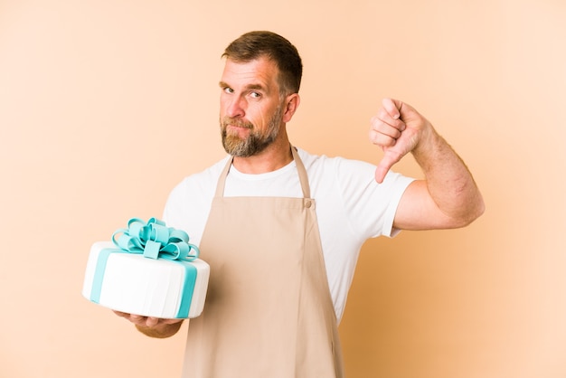 Senior holding a cake isolated on beige background showing a dislike gesture, thumbs down. Disagreement concept.