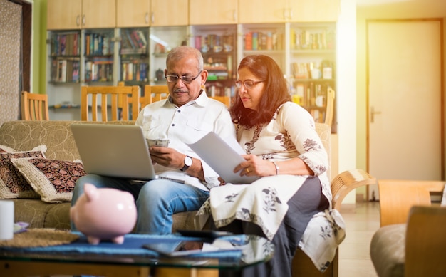 Senior Indian asian couple accounting, doing home finance and checking bills with laptop, calculator and money also with piggy bank while sitting on sofa couch or table at home