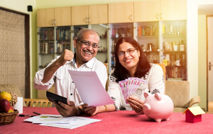 फोटो एक भारतीय एशियाई बुज़ुर्ग दंपति जो घर पर सोफा काउच या टेबल पर बैठे लैपटॉप, कैलकुलेटर और पैसे से होम फाइनेंस का हिसाब कर रहे हैं और बिलों की जाँच कर रहे हैं और साथ ही गुल्लक का भी इस्तेमाल कर रहे हैं।