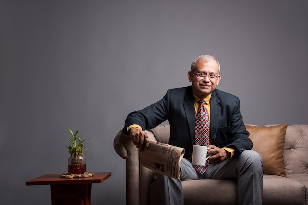 Senior Indian asian executive or businessman in suit and tie, reading news in newspaper or on smartphone while sitting on sofa or couch with a coffee mug