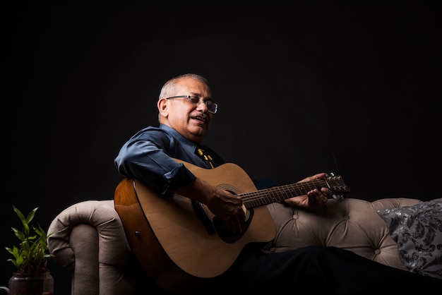 Senior indian asian handsome man playing or posing with musical\
string instrument like guitar or violin, standing over black\
background or sitting on sofa or couch