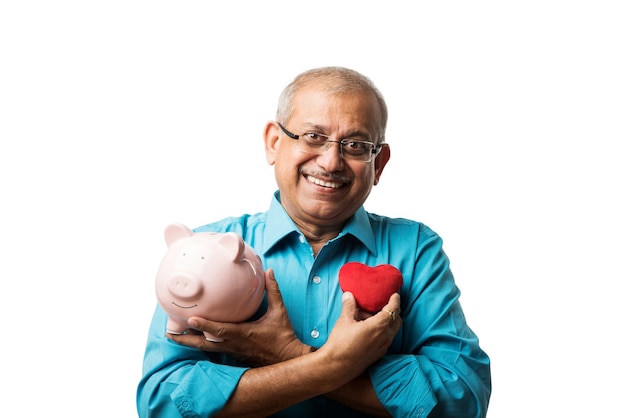 Senior Indian asian man and savings concept - with piggy bank, currency notes, 3d house model, money fan, pile of coins. sitting at table