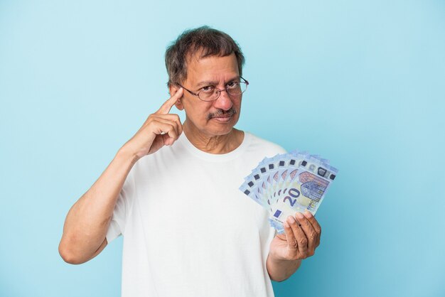 Uomo indiano senior che tiene il conto isolato su sfondo blu che punta il tempio con il dito, pensando, concentrato su un compito.