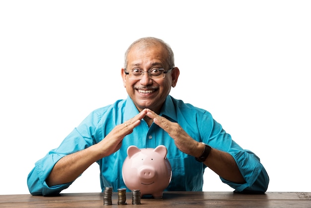 Senior Indiase Aziatische man en spaarconcept - met spaarvarken, bankbiljetten, 3D-huismodel, geldventilator, stapel munten. aan tafel zitten