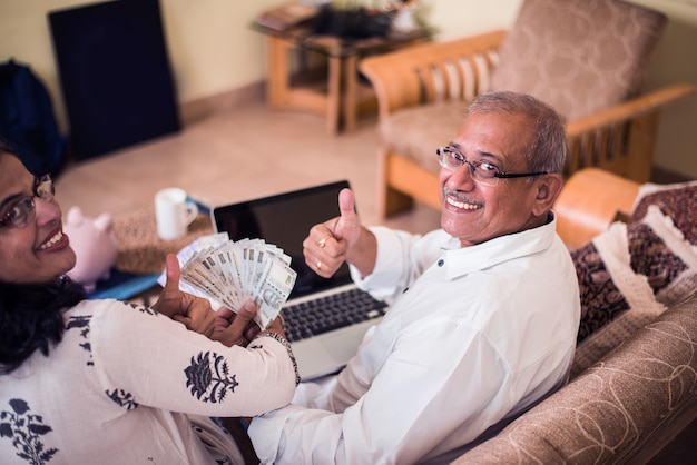 Foto senior indiase aziatische paar boekhouding, huisfinanciering doen en rekeningen controleren met laptop, rekenmachine en geld ook met spaarvarken zittend op de bank of tafel thuis