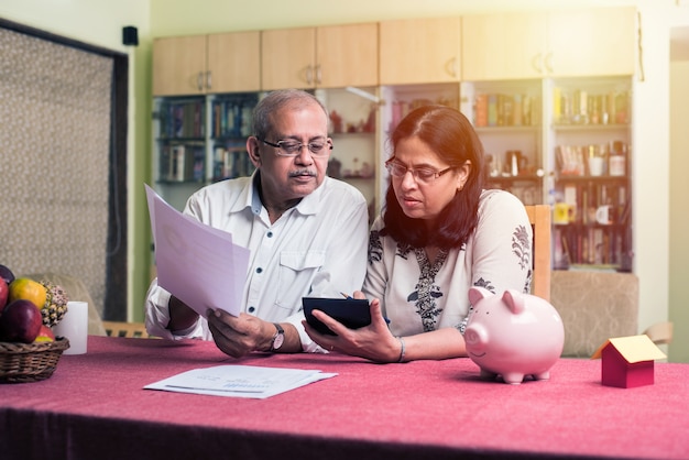 Senior Indiase Aziatische paar boekhouding, huisfinanciering doen en rekeningen controleren met laptop, rekenmachine en geld ook met spaarvarken zittend op de bank of tafel thuis