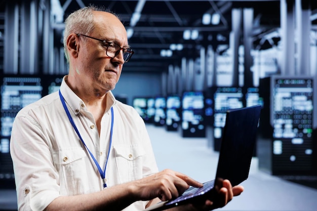 Senior IT developer inspects supercomputers in data center ensuring smooth performance Capable programmer in server farm monitoring energy consumption across units components using laptop