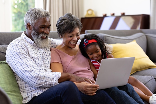 Senior koppel van gemengd ras met hun kleindochter, genietend van hun tijd thuis samen, zittend op een bank, elkaar omhelzend, met behulp van een digitale tablet
