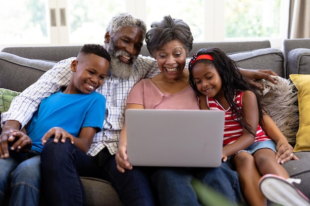 Senior koppel van gemengd ras met hun kleinkinderen, genietend van hun tijd thuis samen, zittend op een bank, elkaar omhelzend, met behulp van een laptop