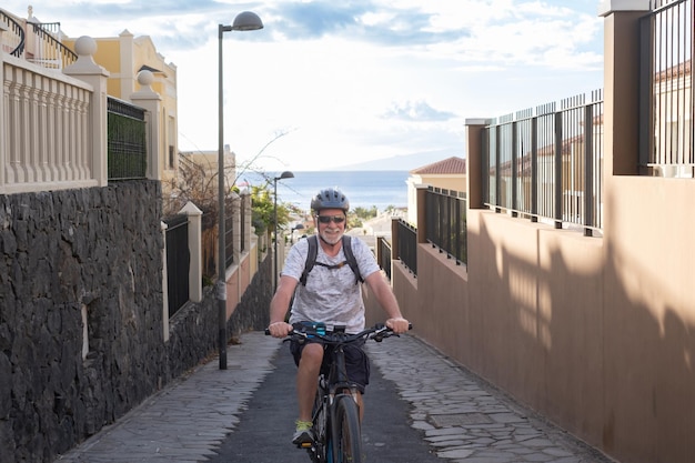 Senior lachende man met helm en zonnebril fietsen met zijn elektrische fiets in bergopwaarts steegje