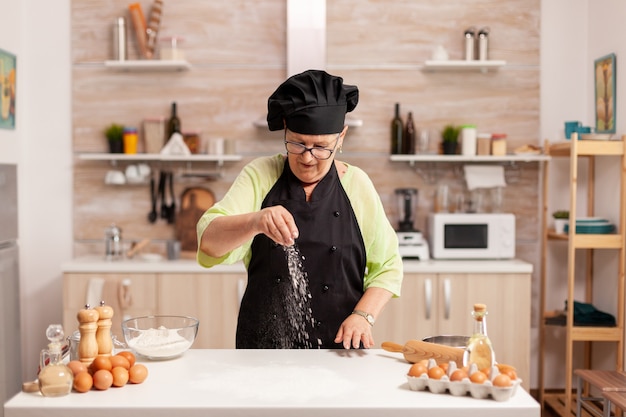 Senior lady chef spreading flour with hand for food preparation in home kitchen wearing apron